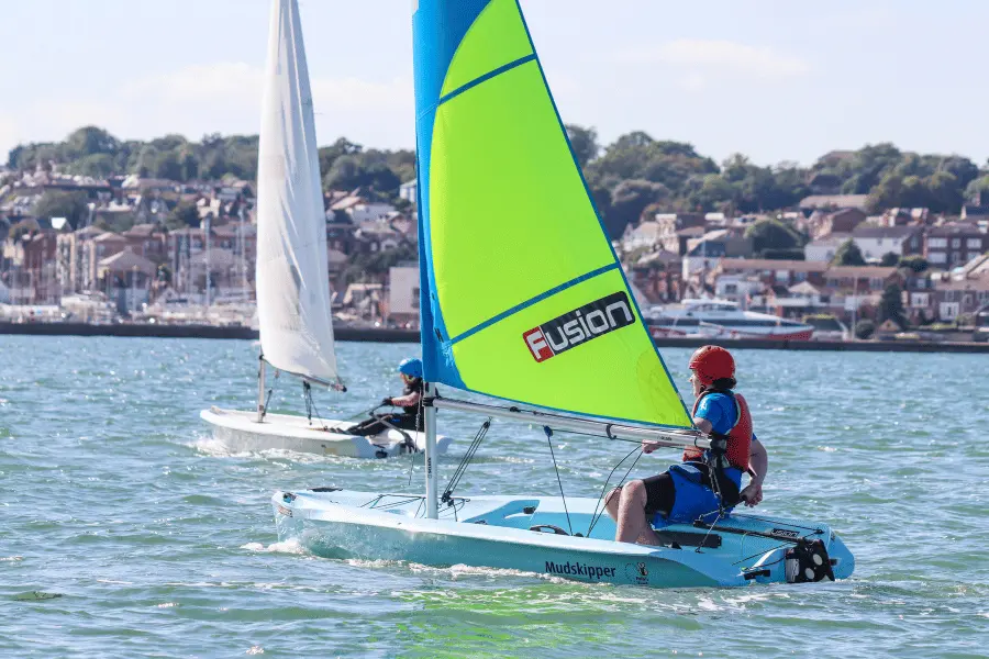 Two students sailing separate small boats with bright blue and green sails on a sunny day, with coastal buildings in the background